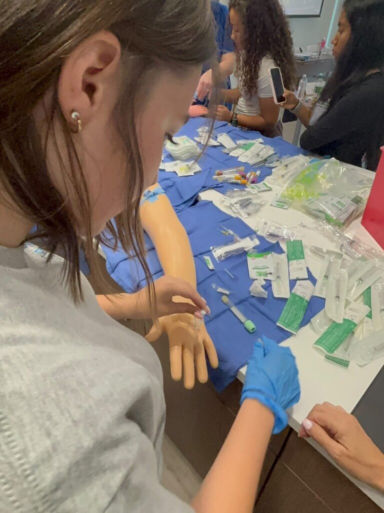 A student practices IV insertion on a realistic training arm placed on a blue‑covered table. Medical supplies such as syringes, alcohol swabs, and packaged equipment are scattered around. Other participants observe in the background.