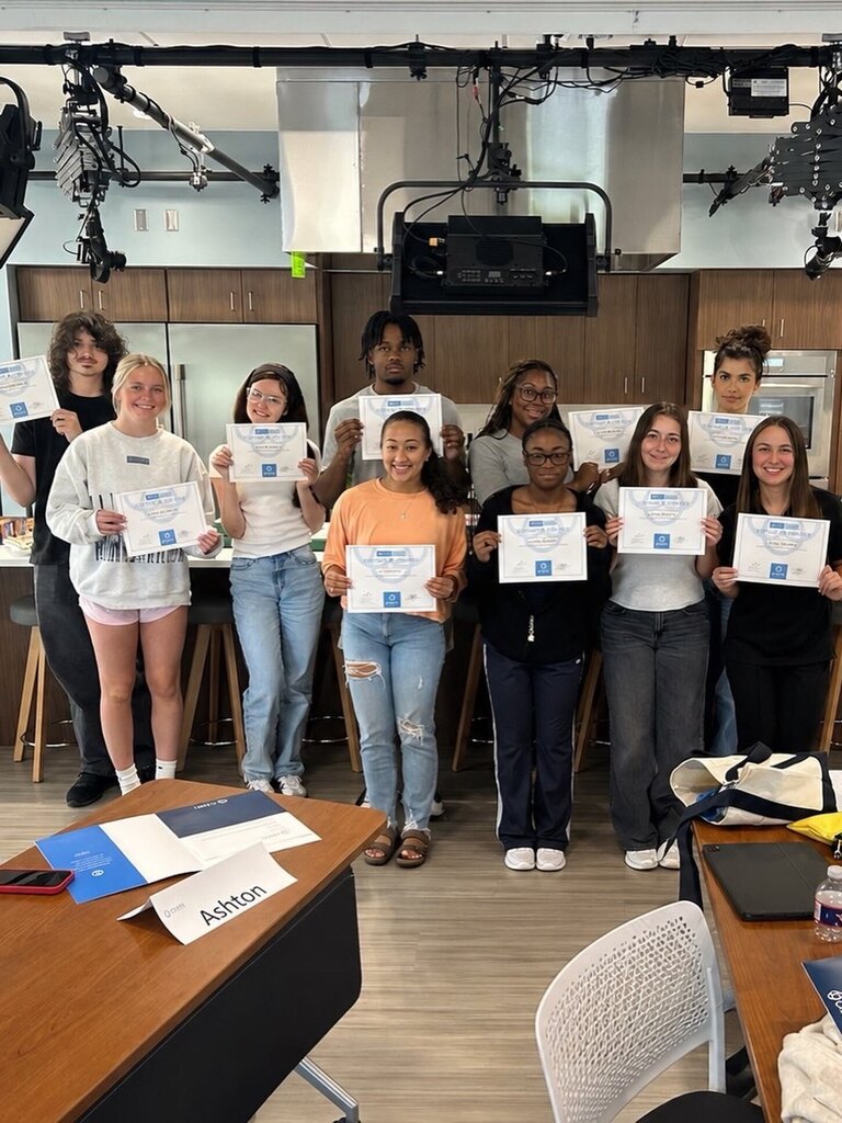 Ten students stand in a kitchen‑style classroom holding certificates. Cabinets, appliances, and overhead lighting equipment fill the background. A table with papers and a name card labeled “Ashton” sits in the foreground.