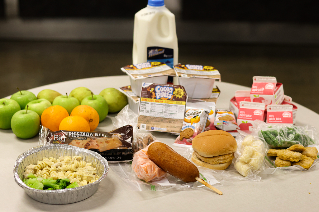 A table displays the full contents of a weekly meal pack, including fresh apples and oranges, a half‑gallon of milk, multiple cartons of apple juice, single‑serve Cocoa Puffs cereal cups, packets of Giant Goldfish cinnamon graham crackers, a foil tray of chicken alfredo with broccoli, a packaged turkey corn dog with carrots, chicken nuggets with green beans, a chicken sandwich with tater tots, and a Fiestada beef stuffed sandwich with refried beans. The items are arranged neatly to show the variety included in the free breakfast and lunch packs for children.