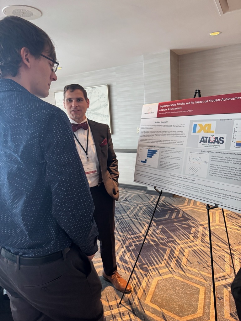 Two individuals stand in front of a research poster titled “Implementation Fidelity and Its Impact on Student Achievement on State Assessments.” One person wears glasses and a blue patterned shirt; the other wears a suit with a red bow tie and a conference badge. The poster behind them includes text sections, charts, and logos for IXL and ATLAS.