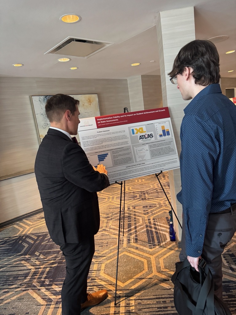 Two individuals stand in a conference setting looking at a research poster on an easel titled “Implementation Fidelity and Its Impact on Student Achievement and Growth on State Assessments.” The poster features text blocks, bar graphs, line charts, and logos for IXL and ATLAS. The room has patterned carpet, neutral walls, and ceiling lights.