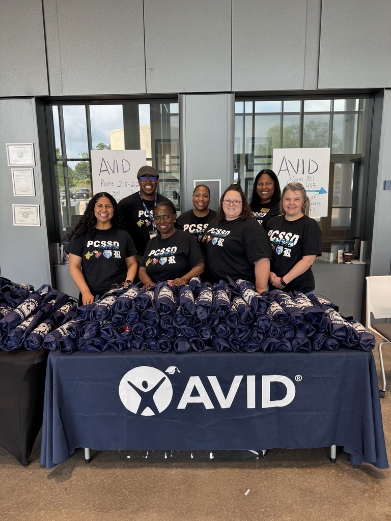 A group of seven staff members stand behind a table covered with rolled AVID materials. They are wearing matching black PCSSD shirts, and signs behind them indicate AVID room locations.