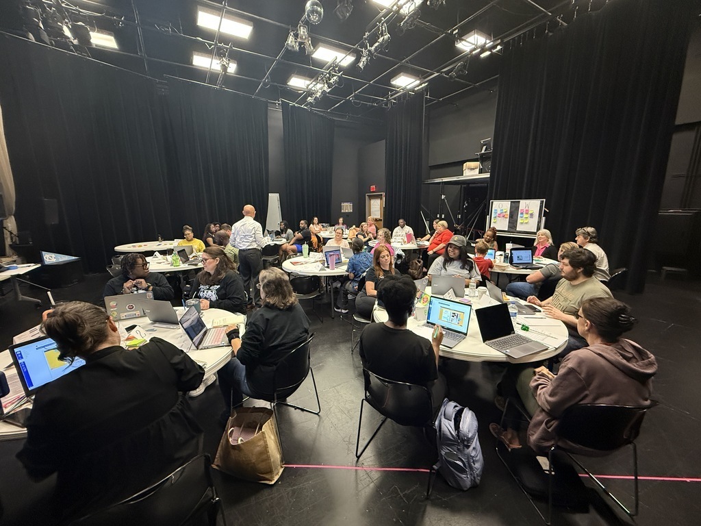 Multiple round tables are filled with educators using laptops and taking notes during a large AVID training session. A facilitator stands near the center addressing the group.