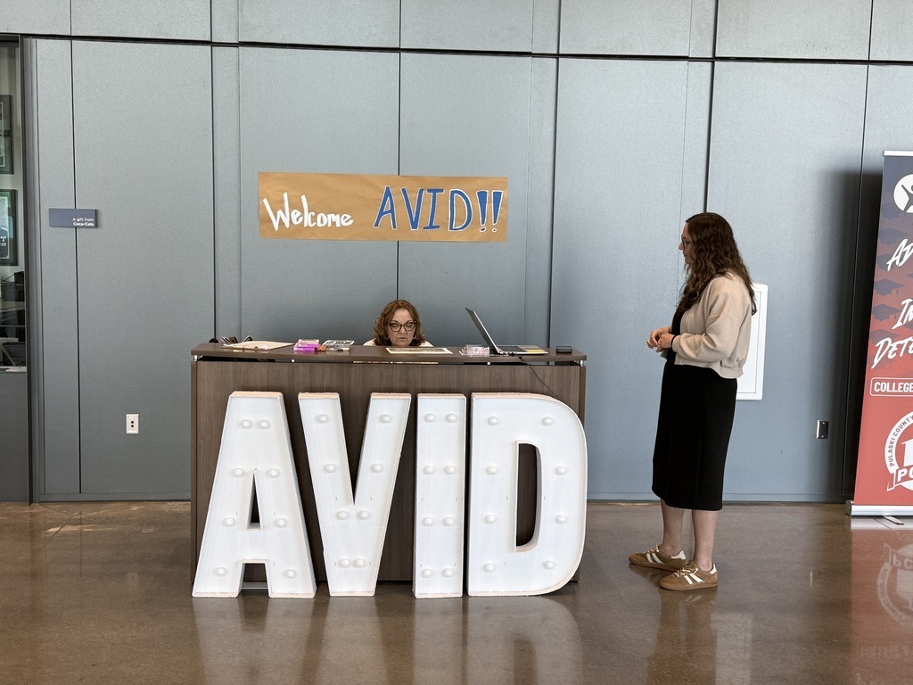 A welcome desk displays large illuminated “AVID” letters. A staff member sits behind the desk working on a laptop while another person stands in front of it. A banner above reads “Welcome AVID!!”