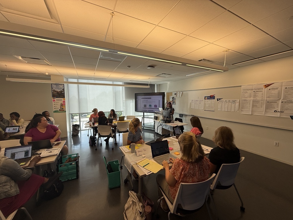 Participants sit at tables with laptops and notebooks during an AVID professional development session. A presenter stands near a screen, and posters about AVID implementation hang on the walls.