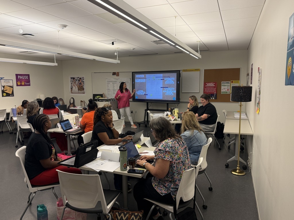 Adults sit at tables with laptops while an instructor in a pink shirt presents at the front of the room. A large screen displays a slide with charts and highlighted sections.