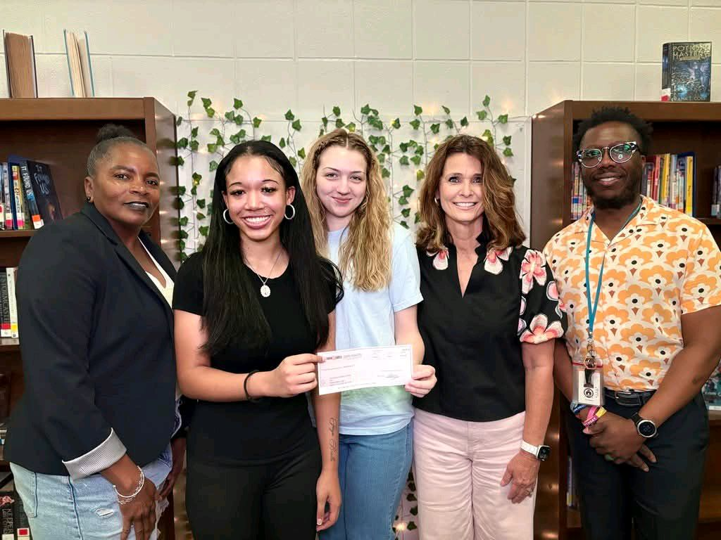 Two students stand side by side in a library space, holding a large scholarship check together. Bookshelves and decorative vines are visible in the background. Both students are smiling as they display the Bobby Agee AVID Scholarship check awarded through Baldwin & Shell and the SHMS PTSA.