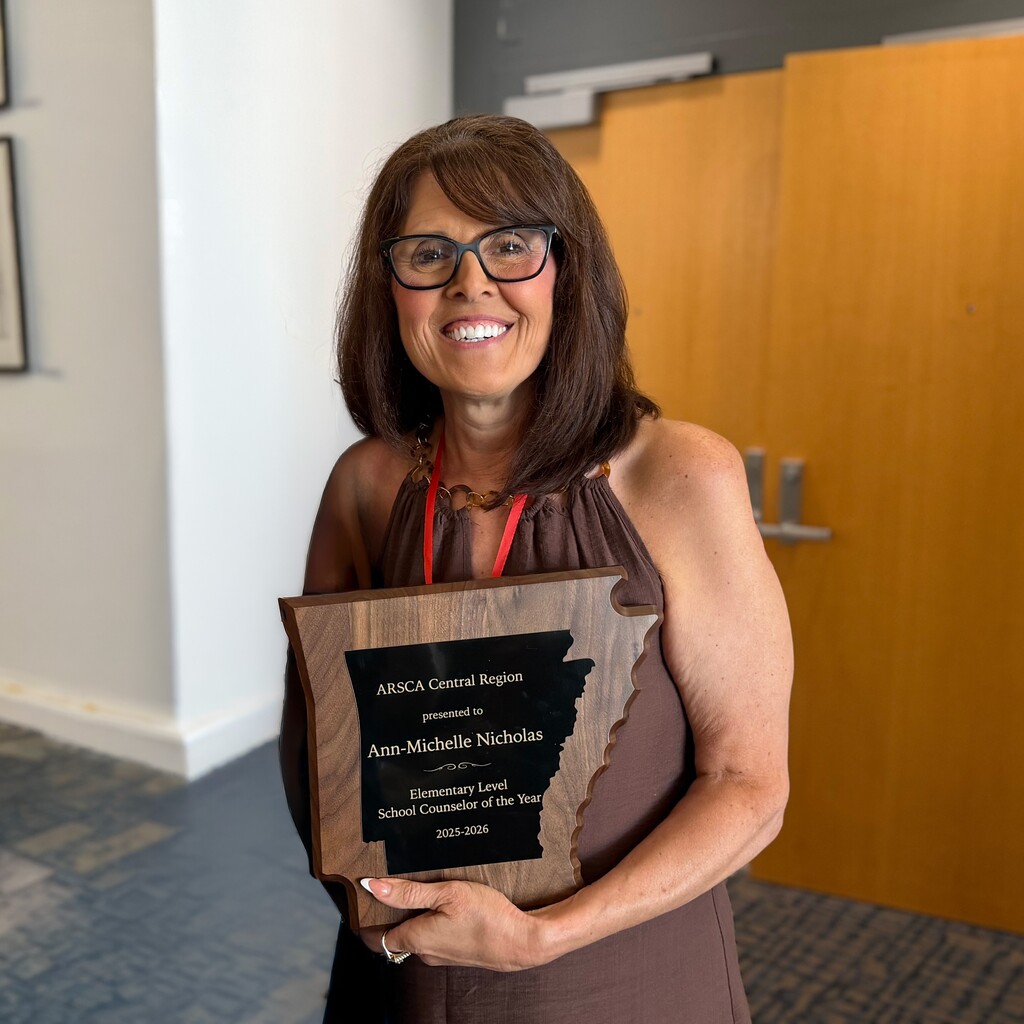 Ann Michelle Nicholas stands indoors holding a wooden Arkansas‑shaped plaque recognizing her as the 2025–2026 ArSCA Central Region Elementary School Counselor of the Year. She is smiling, wearing glasses and a sleeveless brown top, with a wooden door in the background.