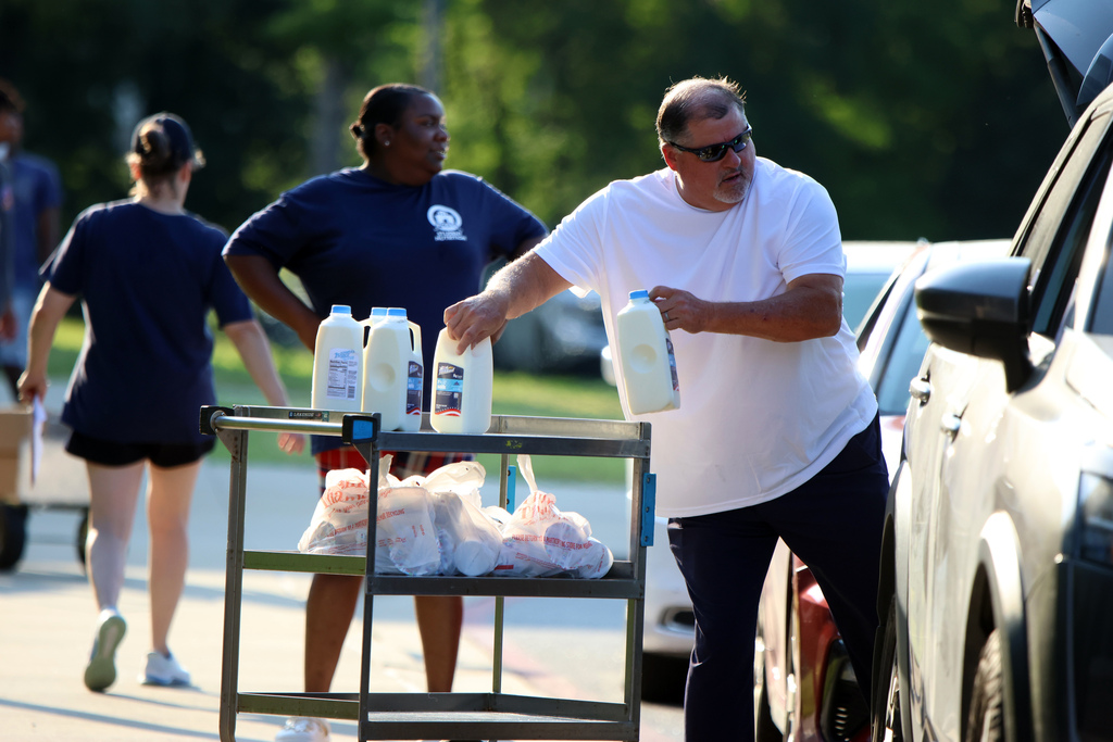 A person in a white shirt and sunglasses places a half-gallon of milk into a vehicle during a food distribution event. A metal cart in front of them holds multiple half-gallons of milk and grocery bags. Two people in dark blue shirts work in the background, one pushing a cart, in a sunny outdoor parking lot.