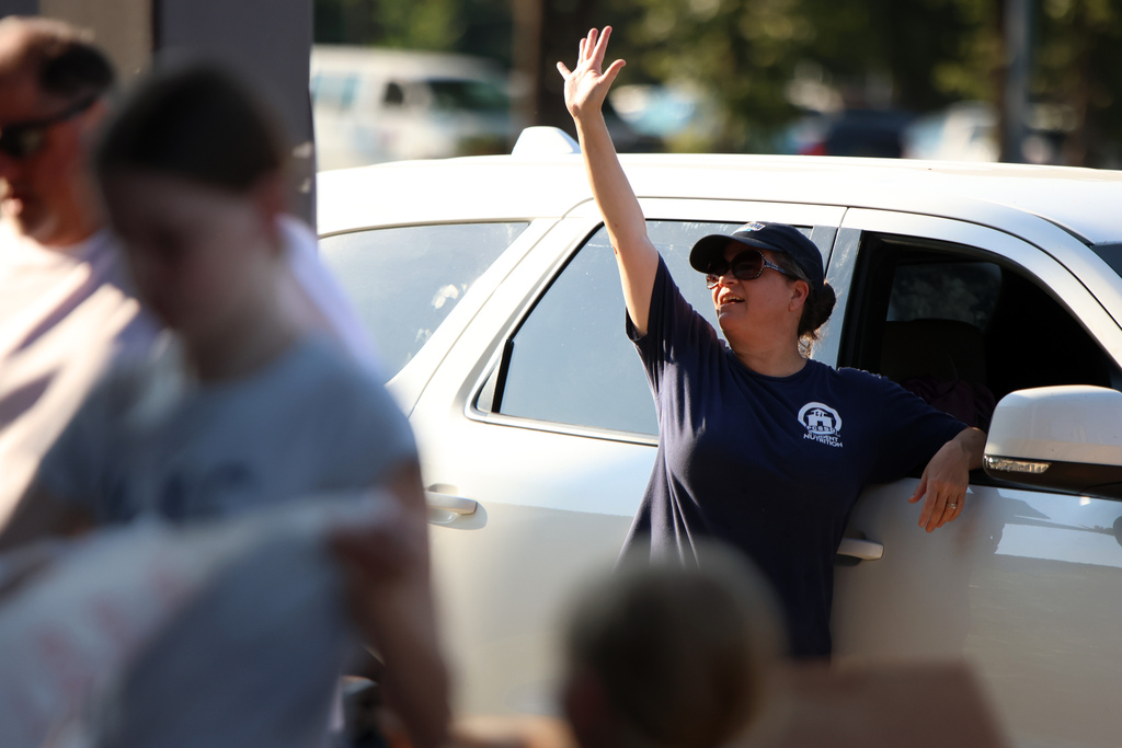 A person wearing sunglasses, a dark cap, and a navy shirt with a “Nutrition” logo stands beside an open vehicle door, leaning on the frame and raising one arm in the air. Other people and vehicles are visible in the background in an outdoor setting.