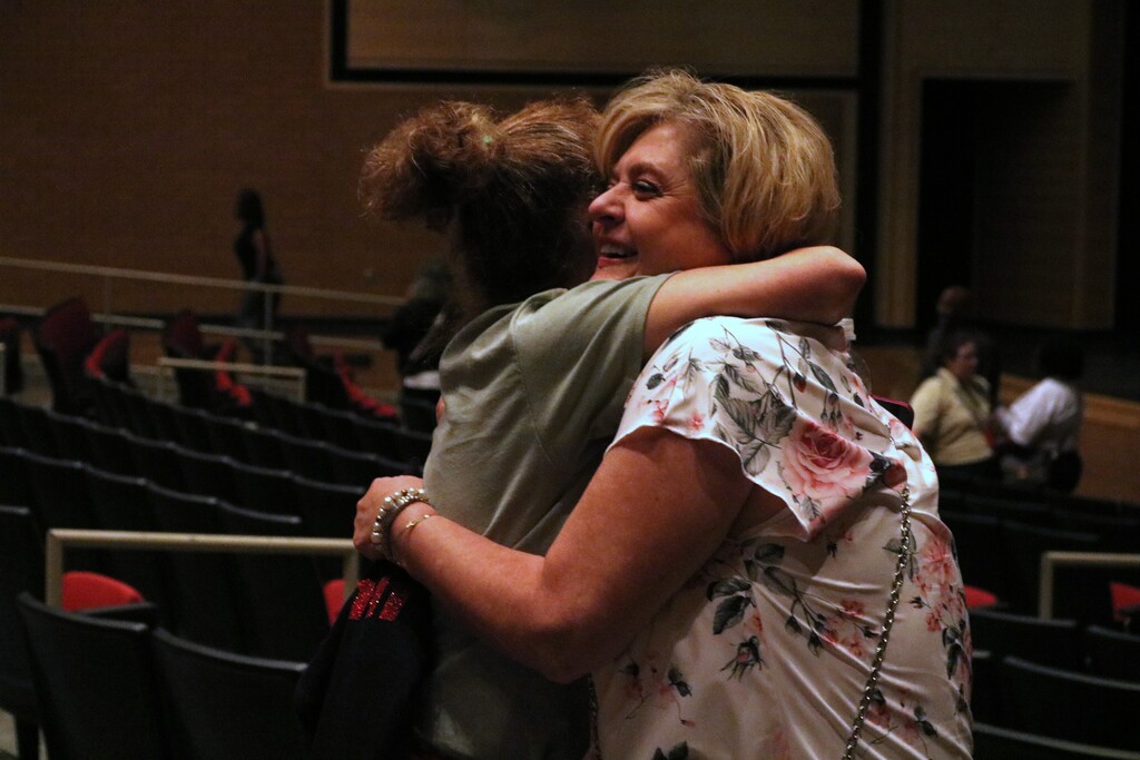 Two people embrace in a hug inside an auditorium, with empty red seats and others talking in the background.