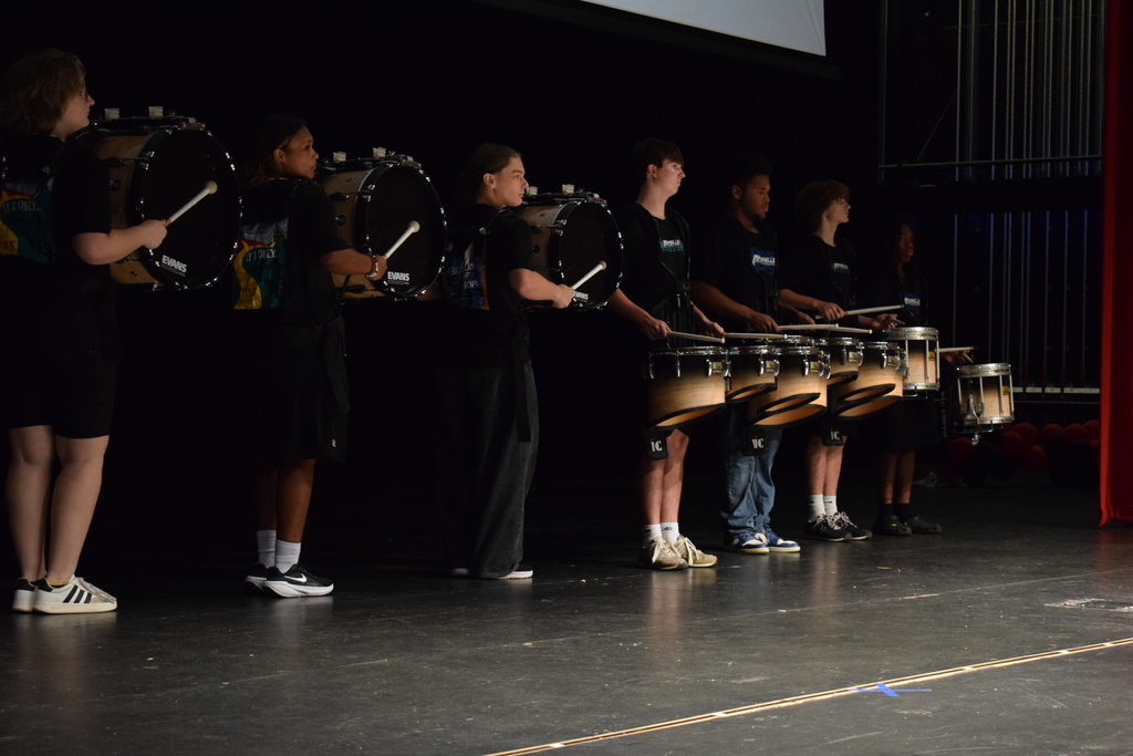 Seven percussionists stand on a stage, each holding bass, snare, or tenor drums in a dimly lit auditorium.
