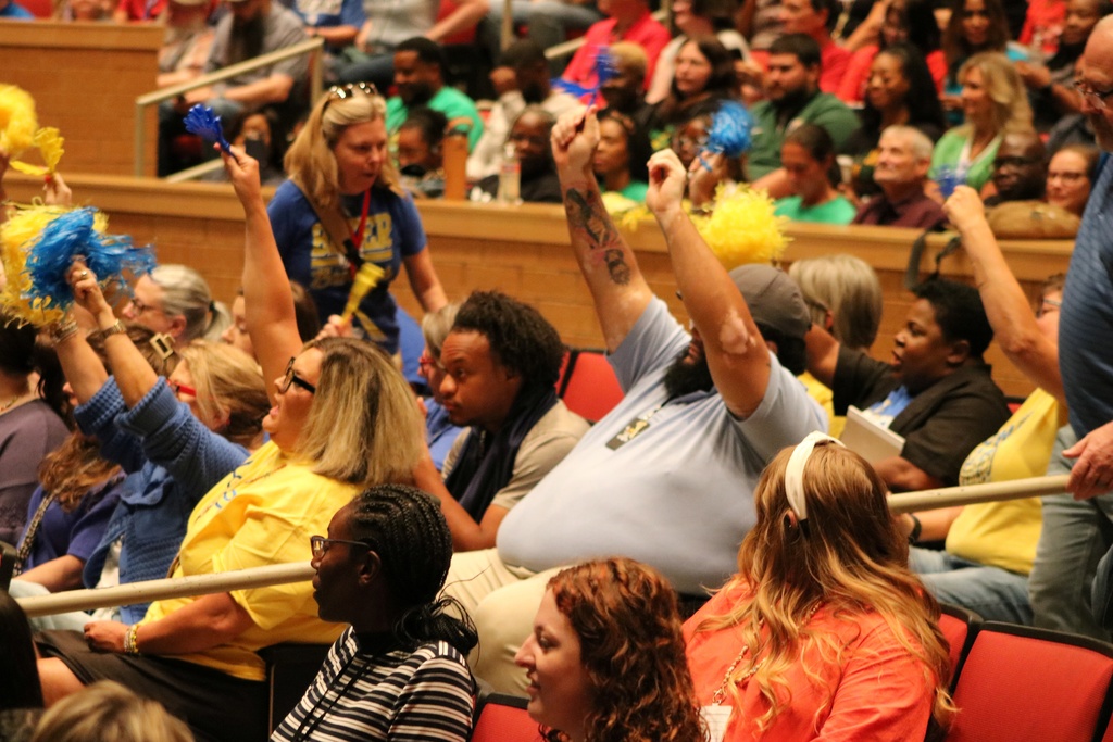 A large audience waves blue and yellow pom‑poms in a tiered auditorium, creating a celebratory scene.