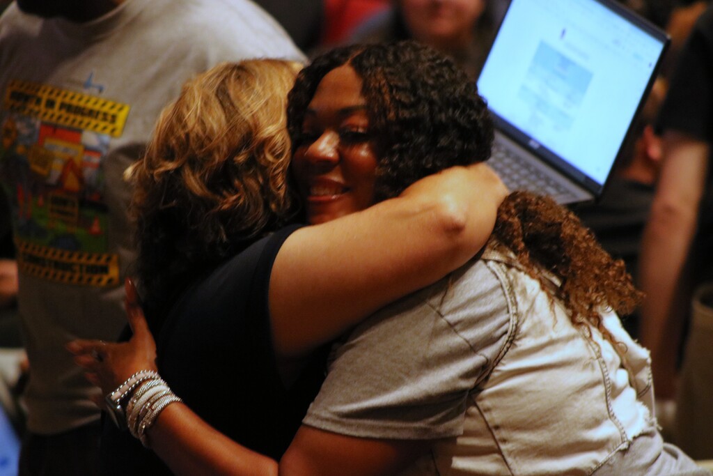  Two people hug closely while others stand nearby; one person in the background holds an open laptop.