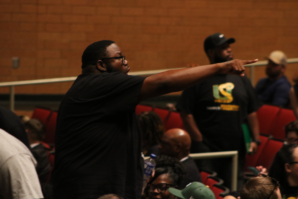 A person stands among seated staff in an auditorium, pointing forward while holding a water bottle.