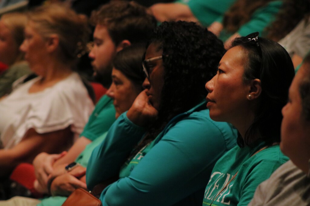 A large group of staff sit closely together in a dim auditorium, facing forward and listening.
