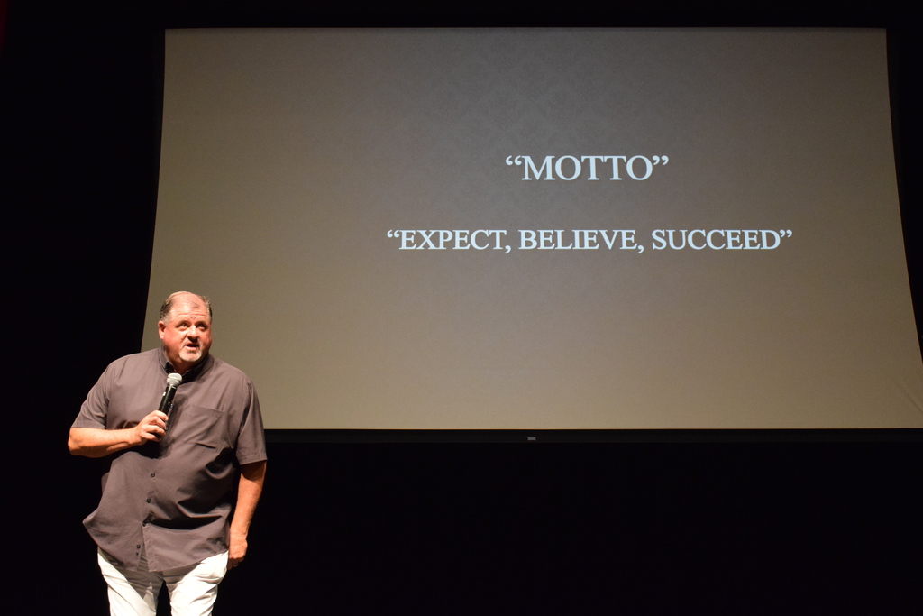 A presenter stands on stage with a projected slide behind them displaying the motto “Expect, Believe, Succeed.”