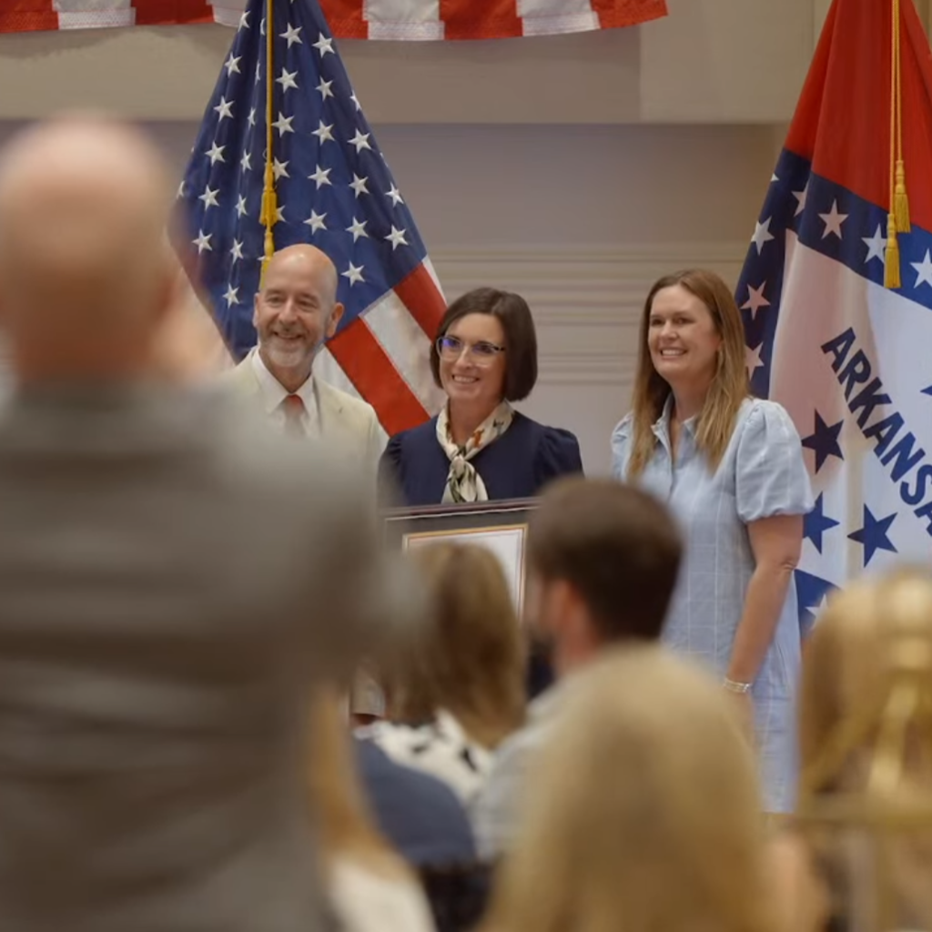 Three people stand together indoors in front of the U.S. and Arkansas flags. The person in the center holds a framed certificate while an audience sits in the foreground.