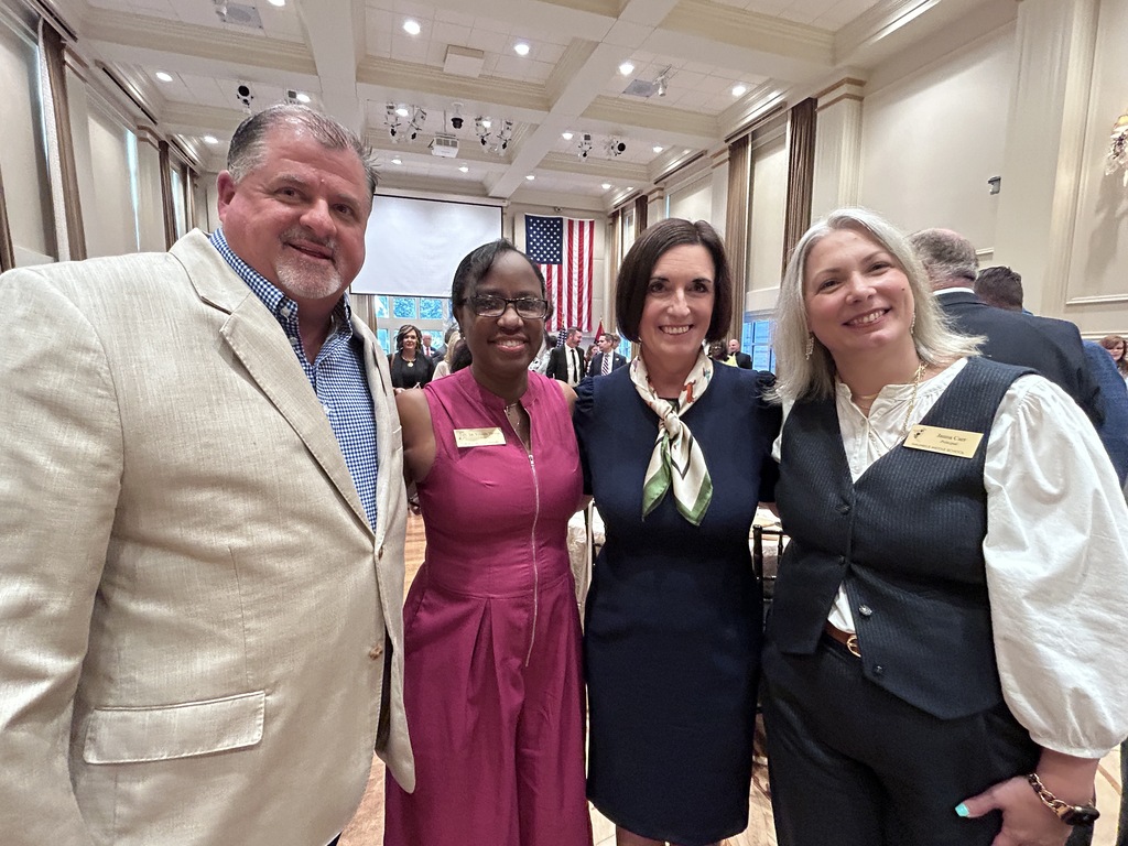 Four people stand together in a formal indoor venue with chandeliers, flags, and a projection screen in the background. Three wear name tags, and other attendees are visible behind them.