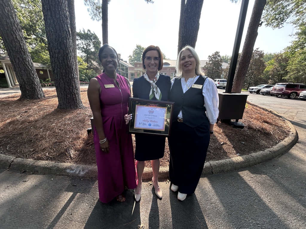 Three people stand outdoors on a paved area surrounded by tall trees. The person in the center holds a framed certificate. A building with columns and parked cars is visible behind them.