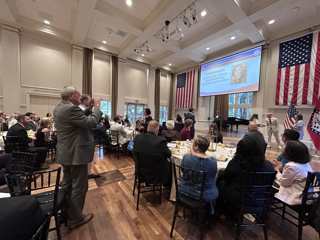 Attendees sit at round tables in a large hall with tall windows and chandeliers. A projection screen at the front displays recognition information for an award finalist. People are standing and applauding.