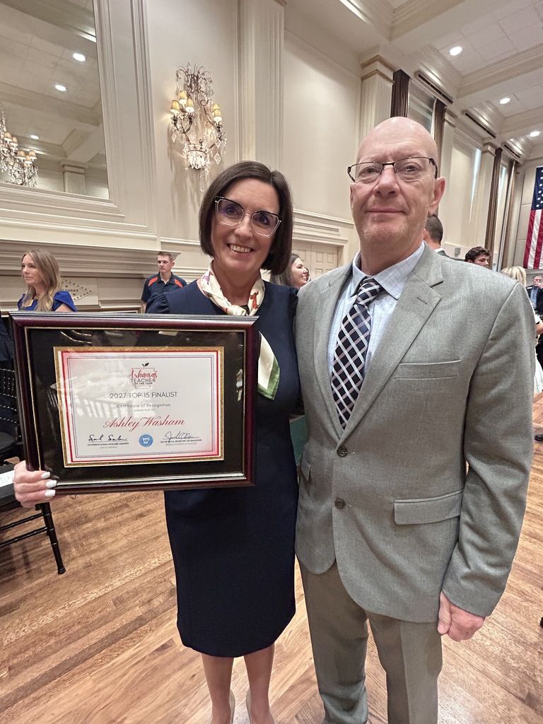 Two people stand together in a formal indoor setting. The person in front holds a framed certificate. Chandeliers, flags, and seated attendees are visible in the background.