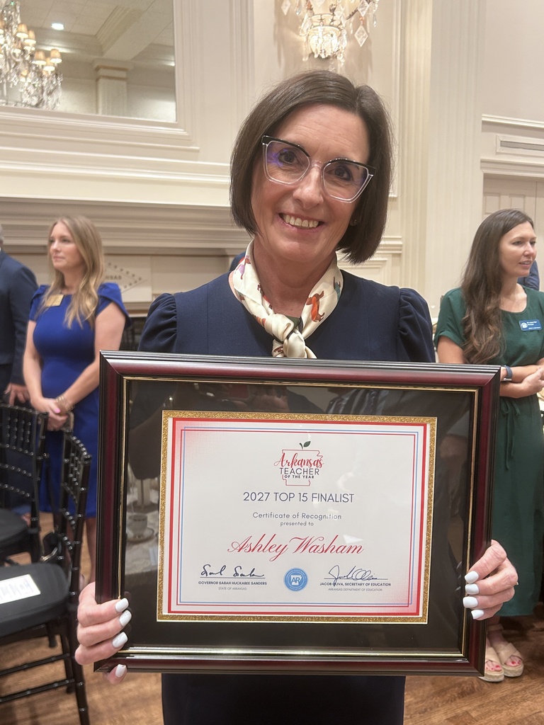 A person stands indoors holding a framed certificate recognizing them as a Top 15 finalist. Other attendees and chandeliers are visible in the background.