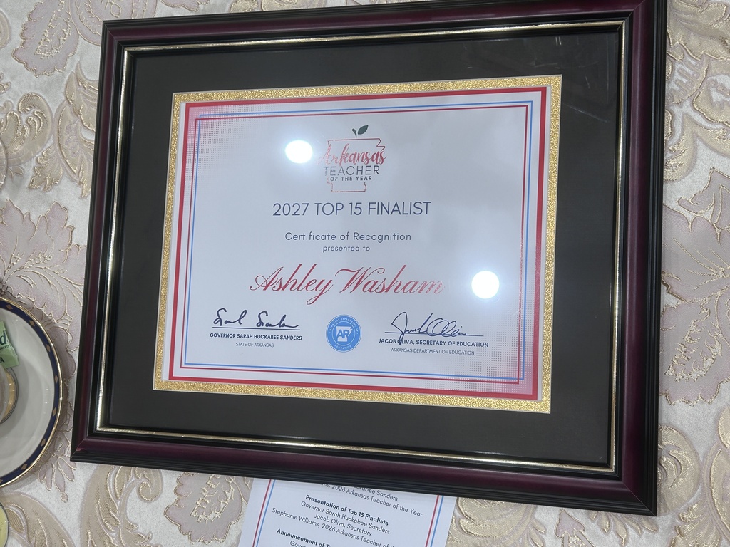 A framed certificate sits on a patterned tablecloth. The certificate recognizes an individual as a Top 15 finalist for a statewide teacher award.