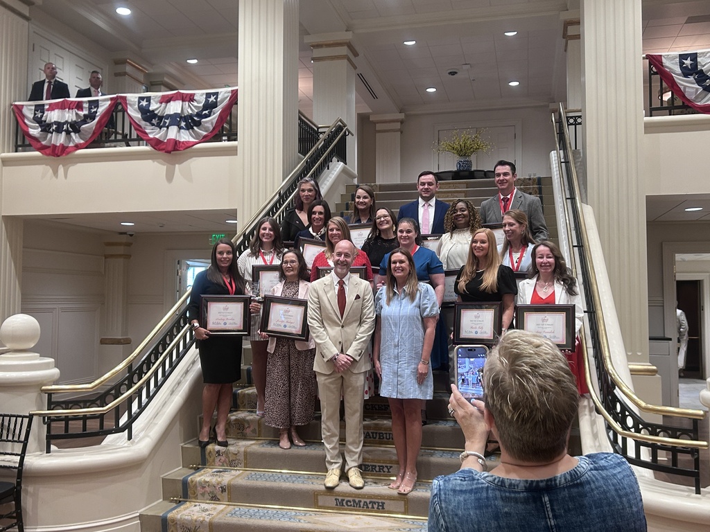 A large group stands on a grand staircase inside a formal building with columns and patriotic bunting. Several people hold framed certificates while someone in the foreground takes a photo.
