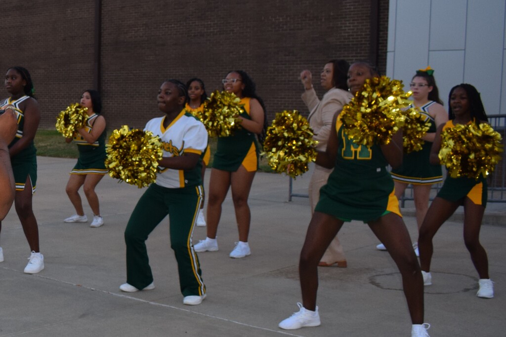 A group of cheerleaders in green and gold uniforms performs with pompoms and enthusiastic expressions.