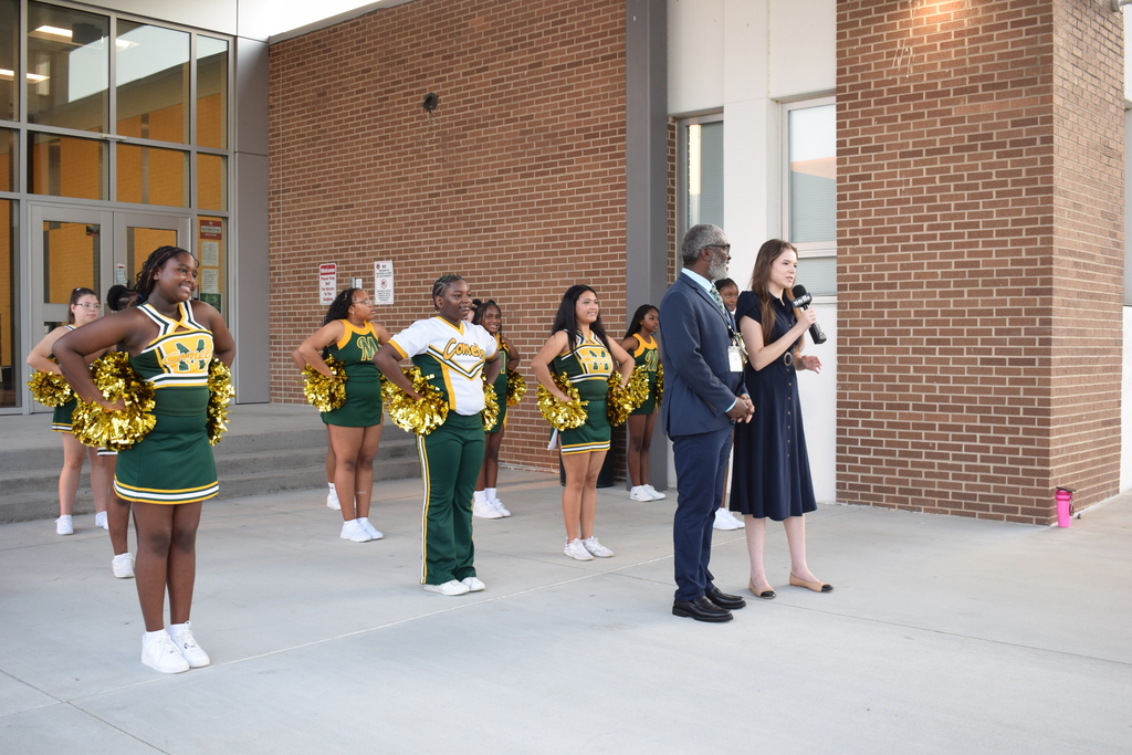 A group of cheerleaders, a reporter and an interviewee stand in front of Mills Middle School.