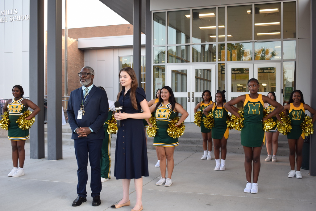 A group of people, including cheerleaders in yellow and green uniforms, standing outside a building with "MILLS MIDDLE SCHOOL" written on it.