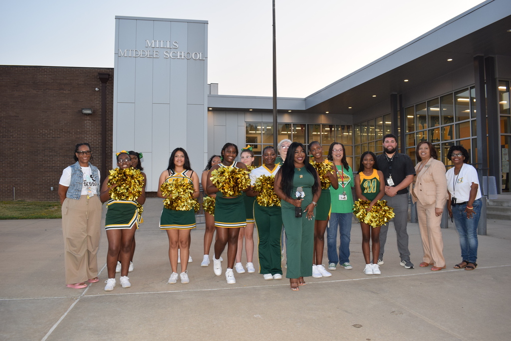 A group of people, including cheerleaders, standing in front of a building with the text "MILLS MIDDLE SCHOOL" visible.