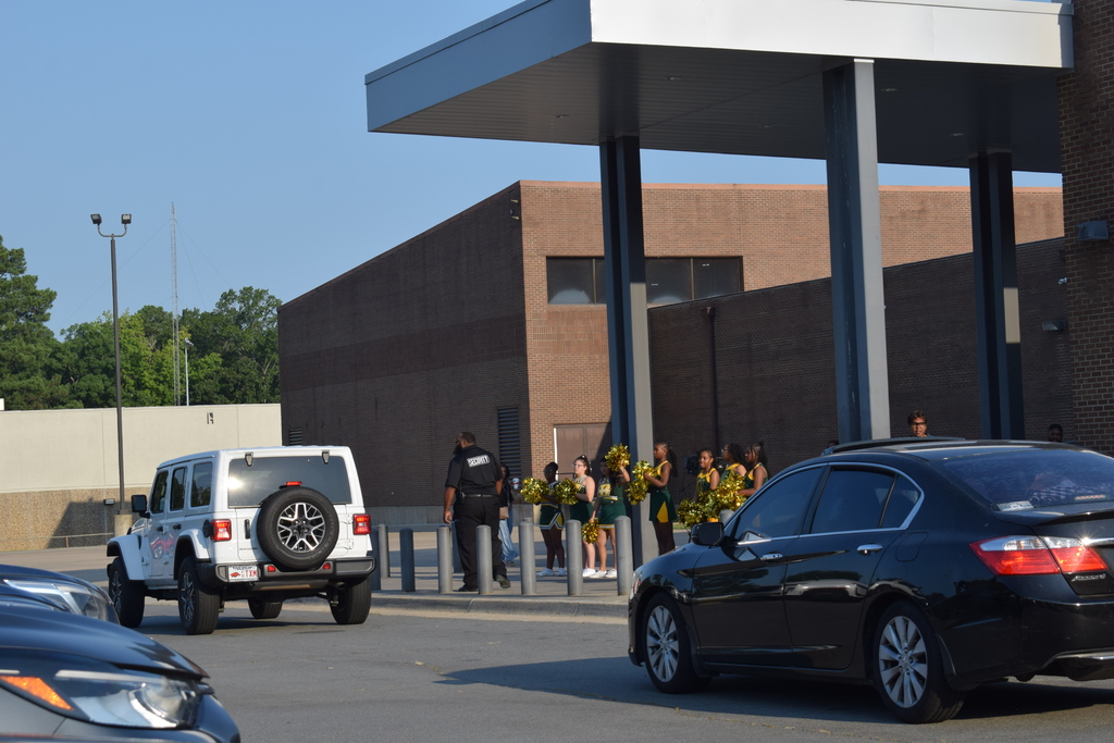 A modern building exterior with an entrance, vehicles parked in front, and several people standing outside.