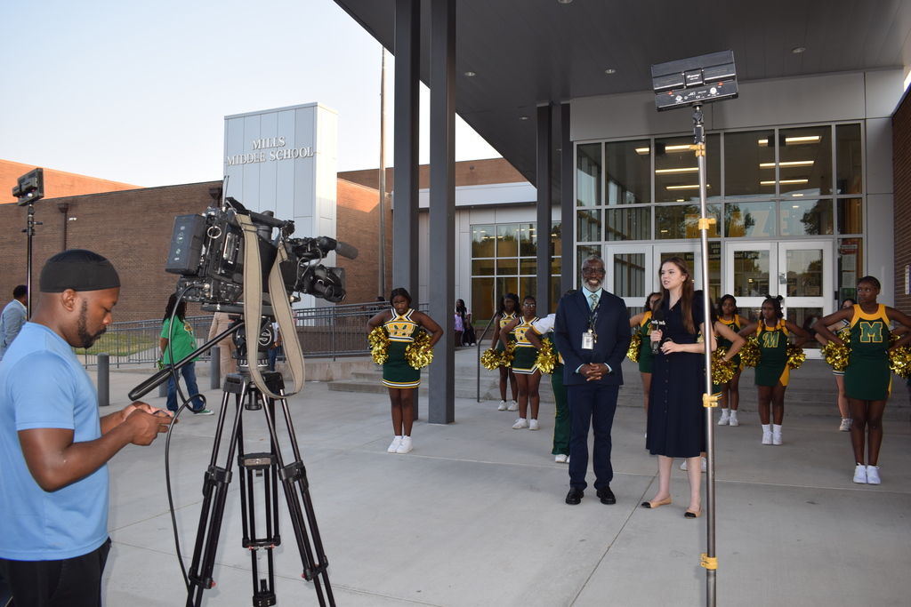 A group of cheerleaders and a cameraman outside a building, with text visible on the building's exterior that says "MILLS MIDDLE SCHOOL"