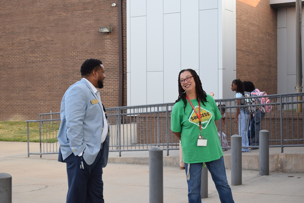 Two people standing outside a brick building, one wearing a green shirt and the other a gray suit, engaged in conversation.