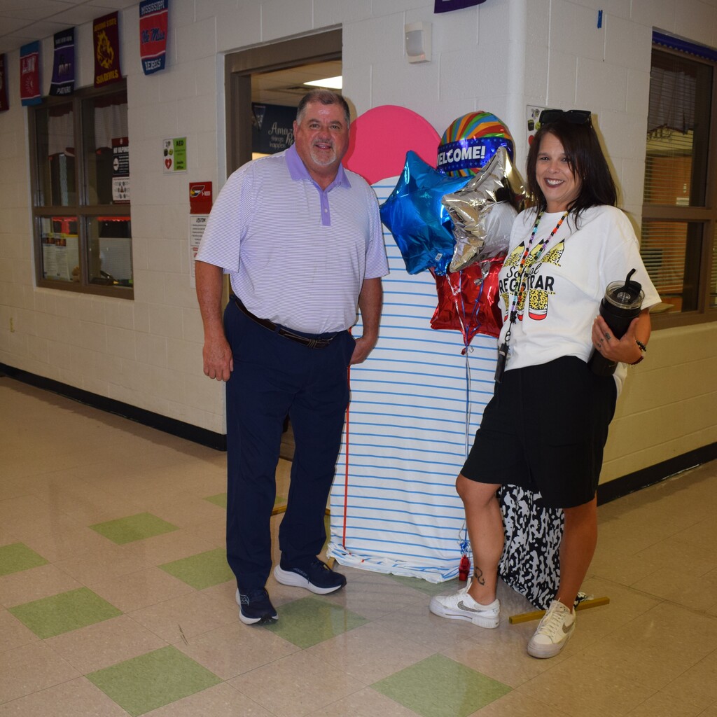 Two adults, a man and a woman, standing in a school hallway with decorations and signage visible.