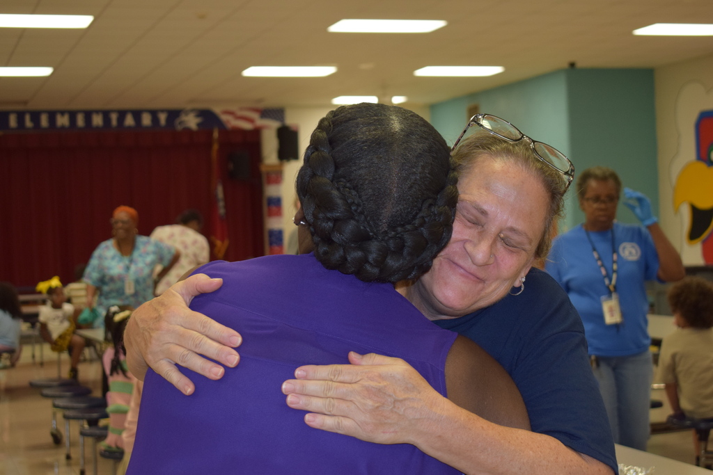 Two people embracing in a school cafeteria, with an "Elementary" sign in the background.