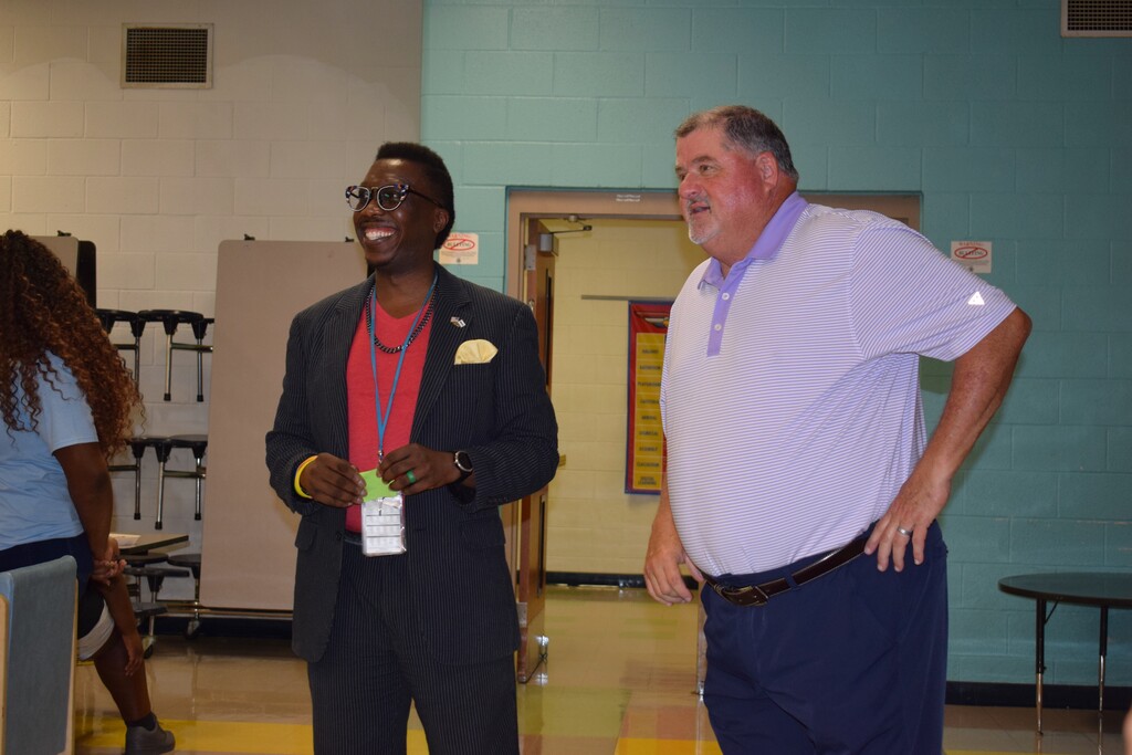 Two men standing and conversing in an indoor setting, one wearing a suit and the other a casual shirt and pants.