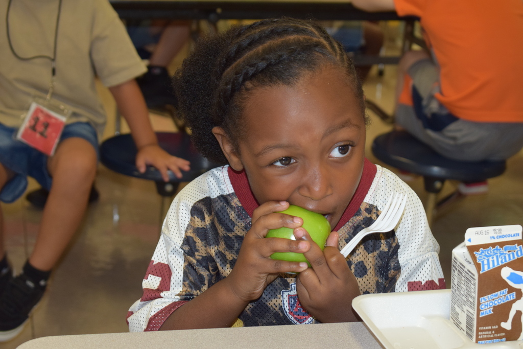Young child with braided hair holding apple and sitting at cafeteria table