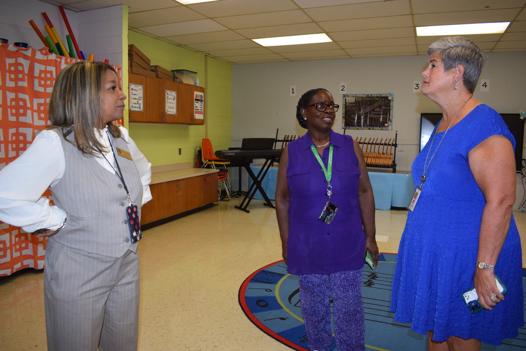 Three women in a classroom, engaged in conversation. The woman on the left wears a gray outfit, the woman in the center wears a purple dress, and the woman on the right wears a blue dress.