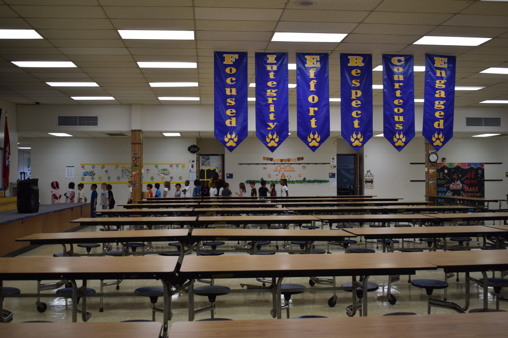 Cafeteria with long tables, blue and gold school banners, and fluorescent lighting fixtures.