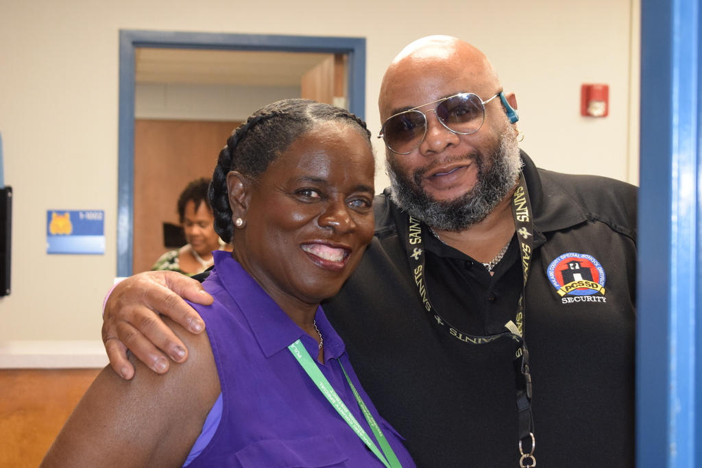 Two smiling adults, one wearing a purple shirt and the other wearing a black shirt with a logo, standing together and embracing.