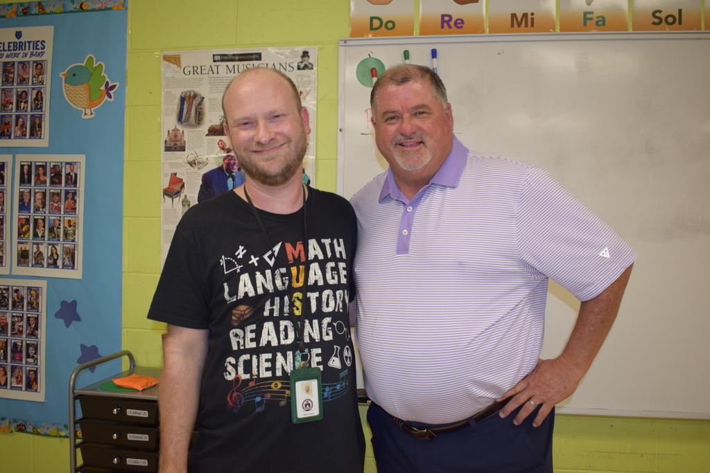 Two men, one wearing a "Music = Math Language History Reading Science" t-shirt, standing in a classroom with educational posters and materials on the walls.