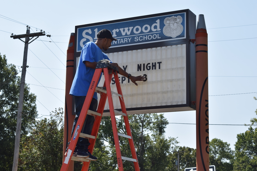 Large outdoor school sign with "Sherwood Elementary School" text and a person on a ladder attaching an event announcement.