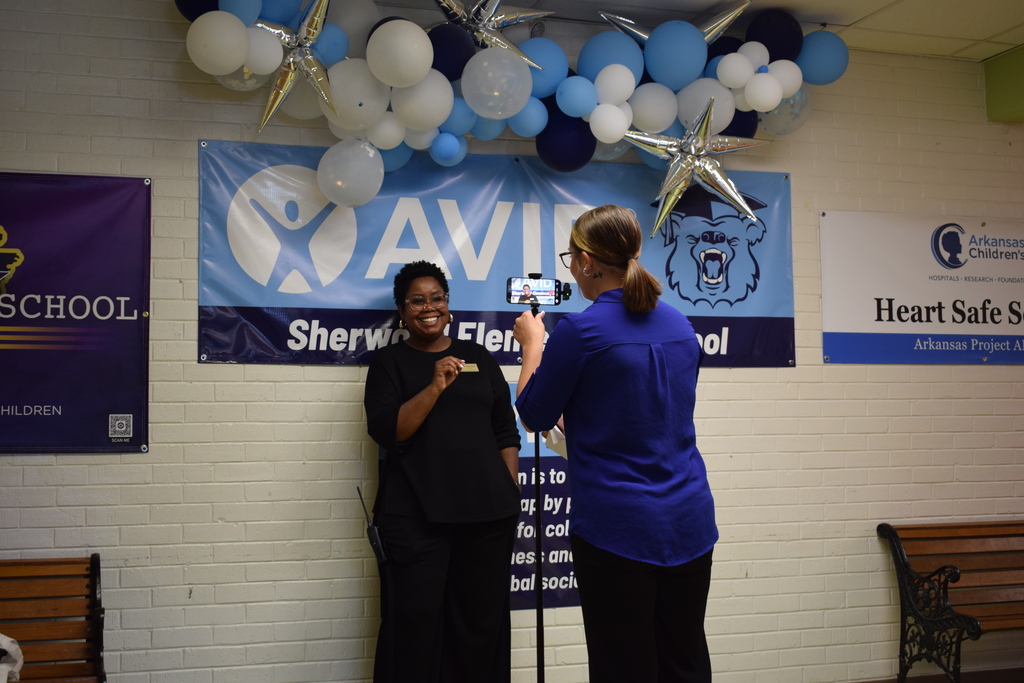 Two people standing in front of a school banner decorated with blue and white balloons.