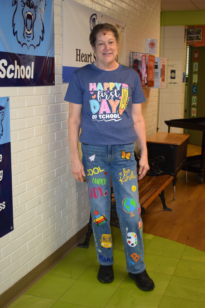 A person wearing a "Happy First Day of School" shirt and pants with school-themed graphics, standing in front of a school hallway.