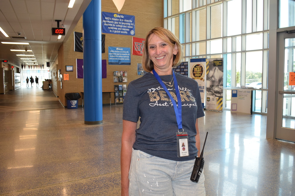A smiling woman wearing a navy blue t-shirt stands in a corridor of a school with signs on the walls.