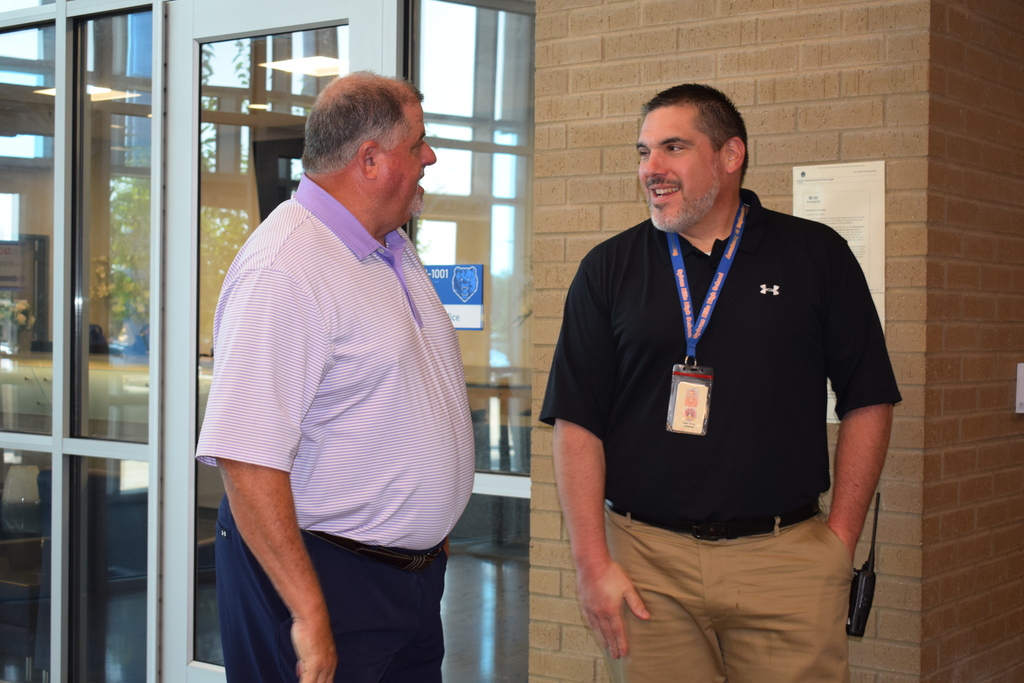 Two middle-aged men in casual clothing conversing in a hallway with windows.
