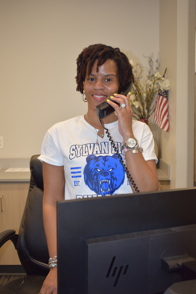 A smiling person wearing a white t-shirt with a blue bear graphic, sitting at a computer and talking on a phone.